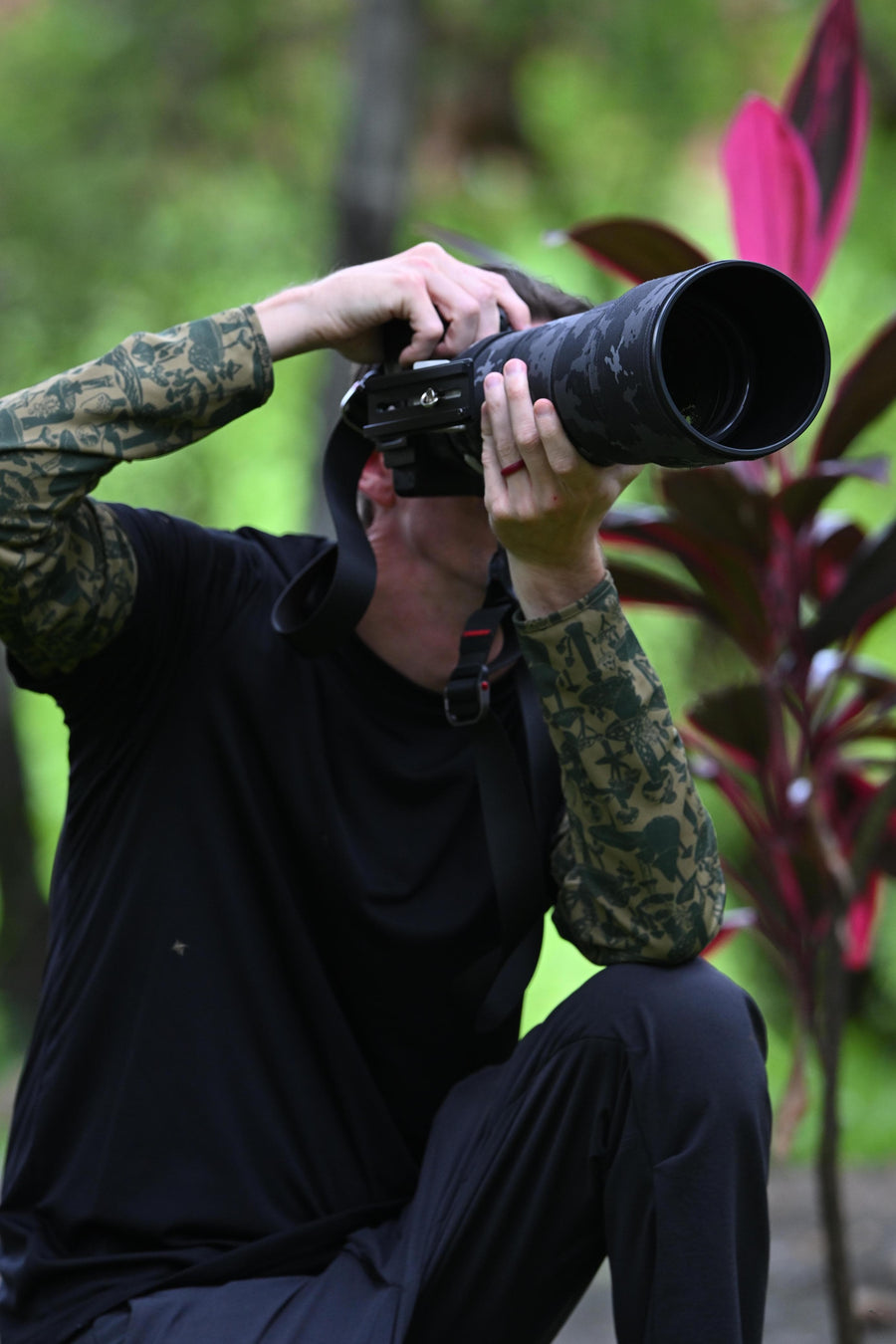 A person crouching outdoors in a black shirt and camouflage sleeves, holding a large black telephoto lens to their eye like a binocular. The background is a blurred green garden with red-leafed plants.